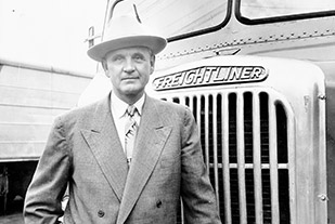Leland James standing proudly in front of an original Freightliner truck, captured in a vintage photograph.
